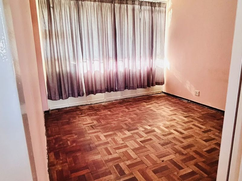 An empty bedroom featuring traditional parquet flooring and a large window with curtains, offering a blank canvas for renovation or interior design.