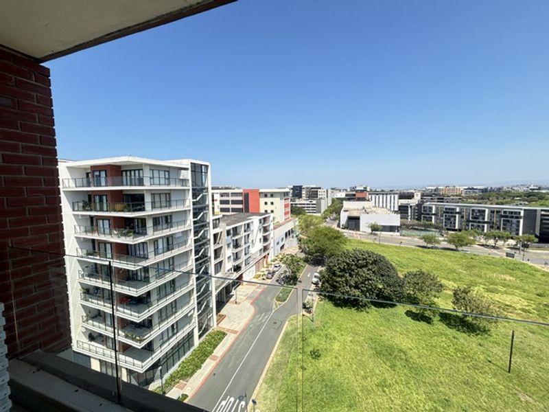 Elevated urban view overlooking surrounding apartment complexes, green space, and local street infrastructure from a balcony with a modern glass balustrade.