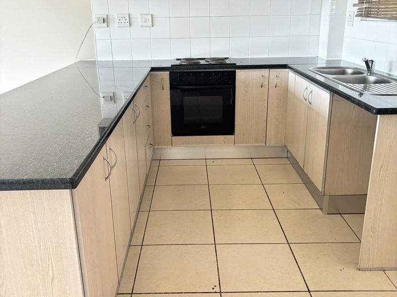 A functional kitchen featuring light wood-finish cabinetry, granite countertops, built-in oven with hob, and a double stainless steel sink with tiled flooring.