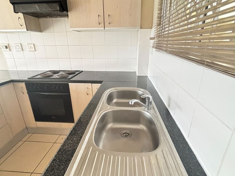 A functional kitchen featuring light wood-finish cabinetry, dark laminate countertops, a double stainless steel sink, and integrated electric cooking appliances with tiled splashbacks.