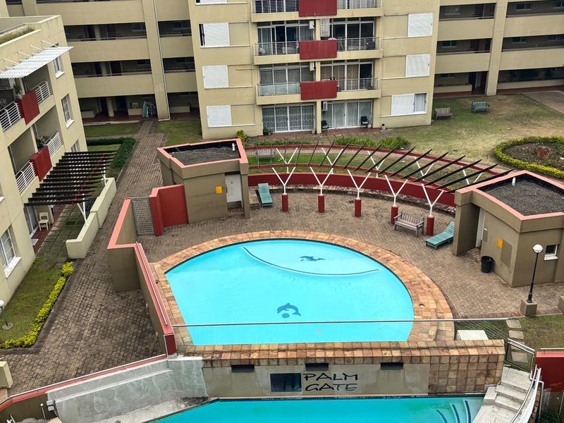 Aerial view of a communal swimming pool area within an apartment complex, featuring paved surrounds, a pergola, and landscaped garden spaces.