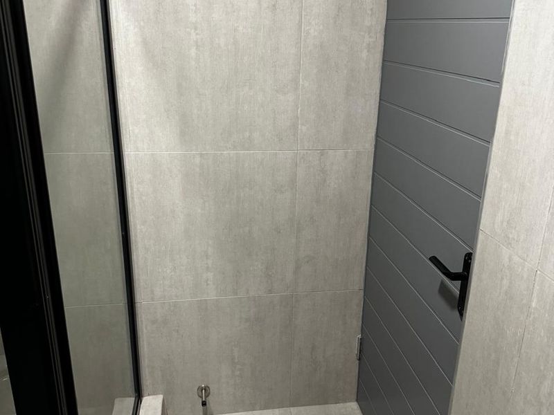 A modern bathroom featuring grey tiled walls and flooring, a sleek grey panelled door with black hardware, and a black-framed shower enclosure.