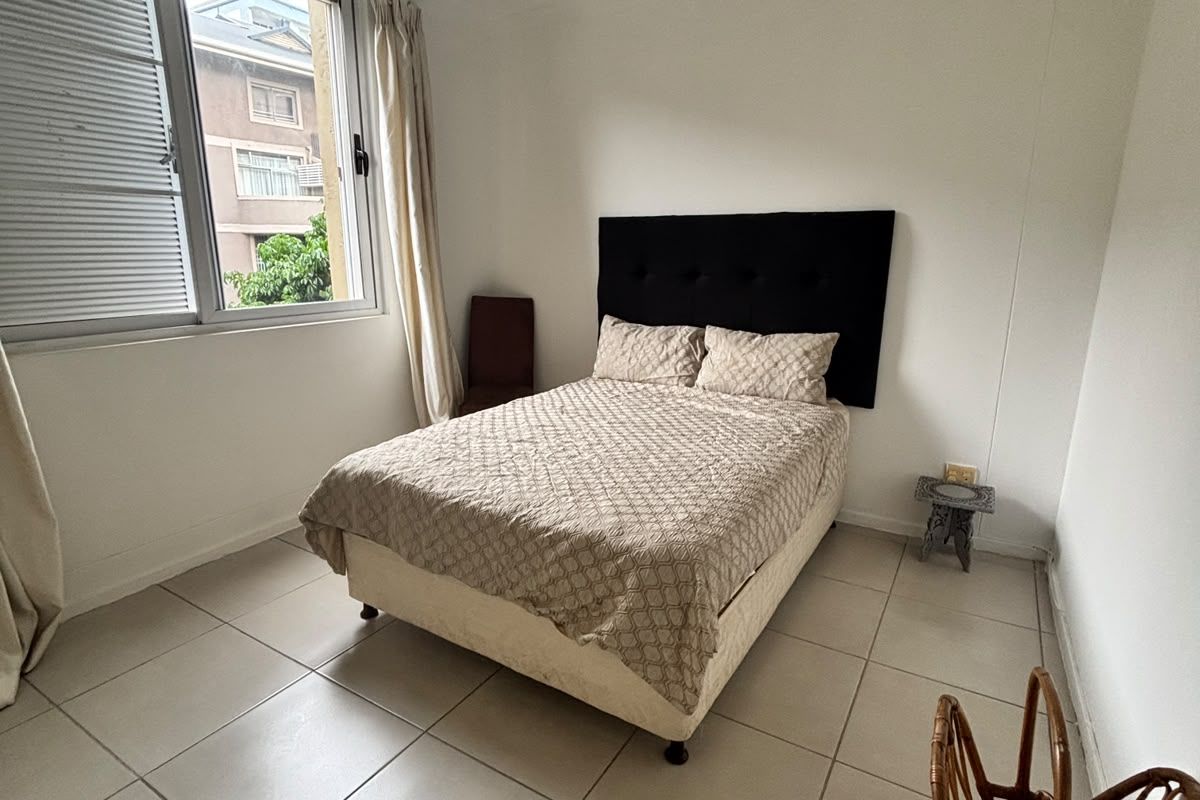 A simple bedroom featuring neutral tiled flooring, a large window with aluminium frames, and a ceiling light fixture.