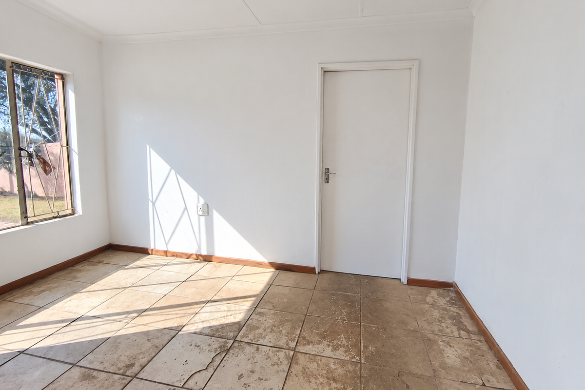 An empty bedroom featuring tiled flooring with visible wear and cracks, white walls, and a window fitted with security bars.