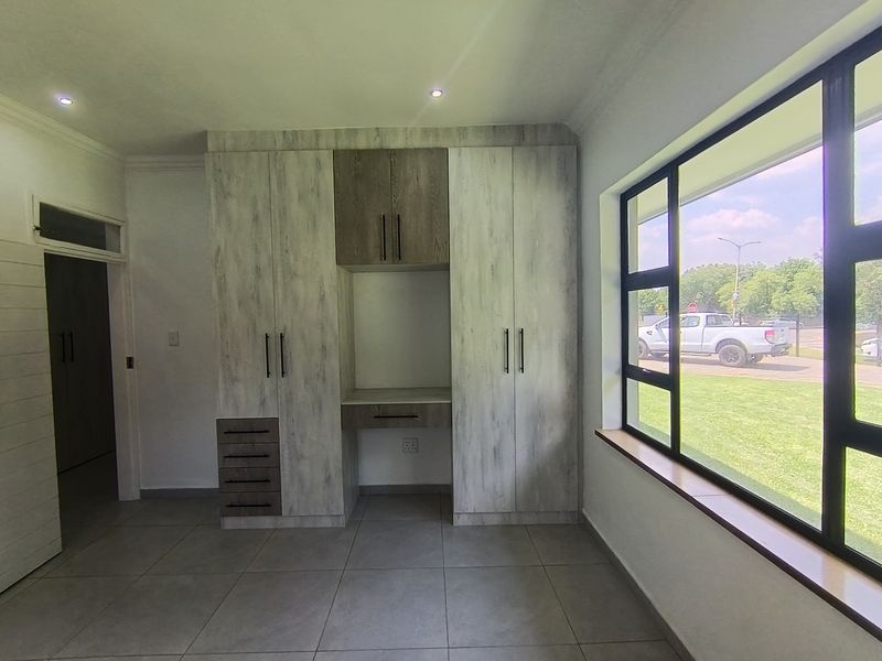 A bright, modern bedroom featuring extensive built-in cabinetry with an integrated desk, large aluminium-framed windows, and neutral tiled flooring.