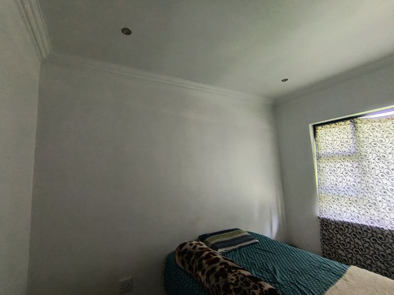 A simple bedroom featuring recessed downlighting, decorative ceiling cornicing, and a window with patterned blinds.