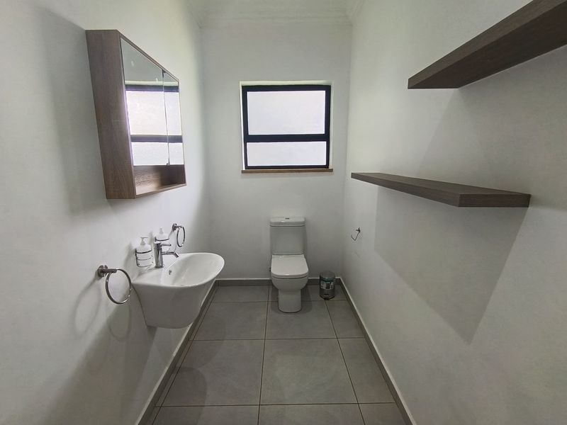 A clean and modern bathroom featuring a wall-mounted basin, toilet, medicine cabinet, and floating shelving with neutral floor tiles.