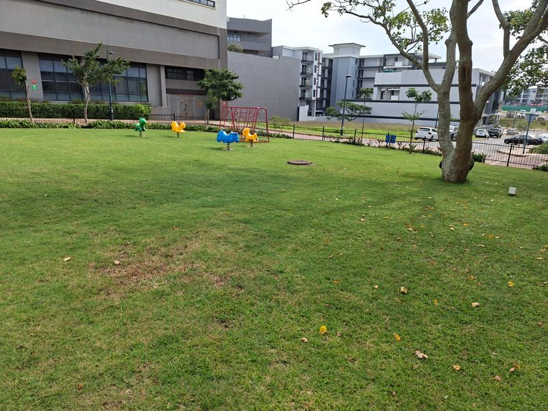 A well-maintained communal garden area featuring a large lawn, mature trees, and perimeter fencing within an apartment complex setting.