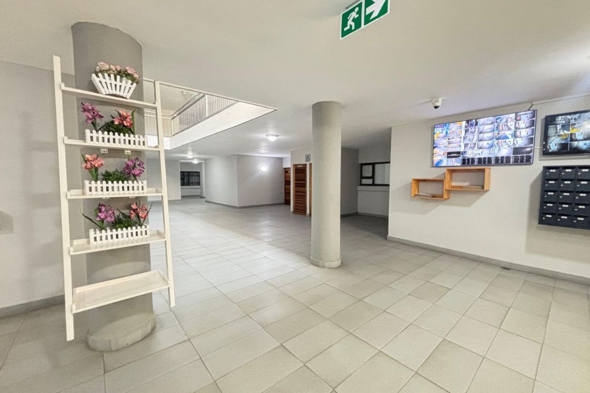 A spacious, well-maintained apartment building lobby or hallway featuring tiled flooring, structural columns, security monitoring screens, and wall-mounted mailboxes.