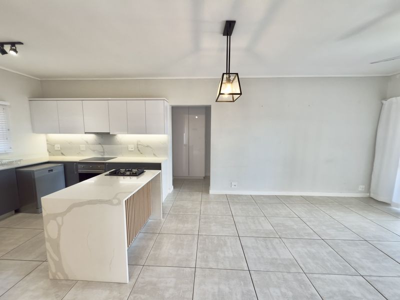 Modern open-plan kitchen and living area featuring a central island with quartz countertops, sleek cabinetry, built-in appliances, and neutral tiled flooring throughout.