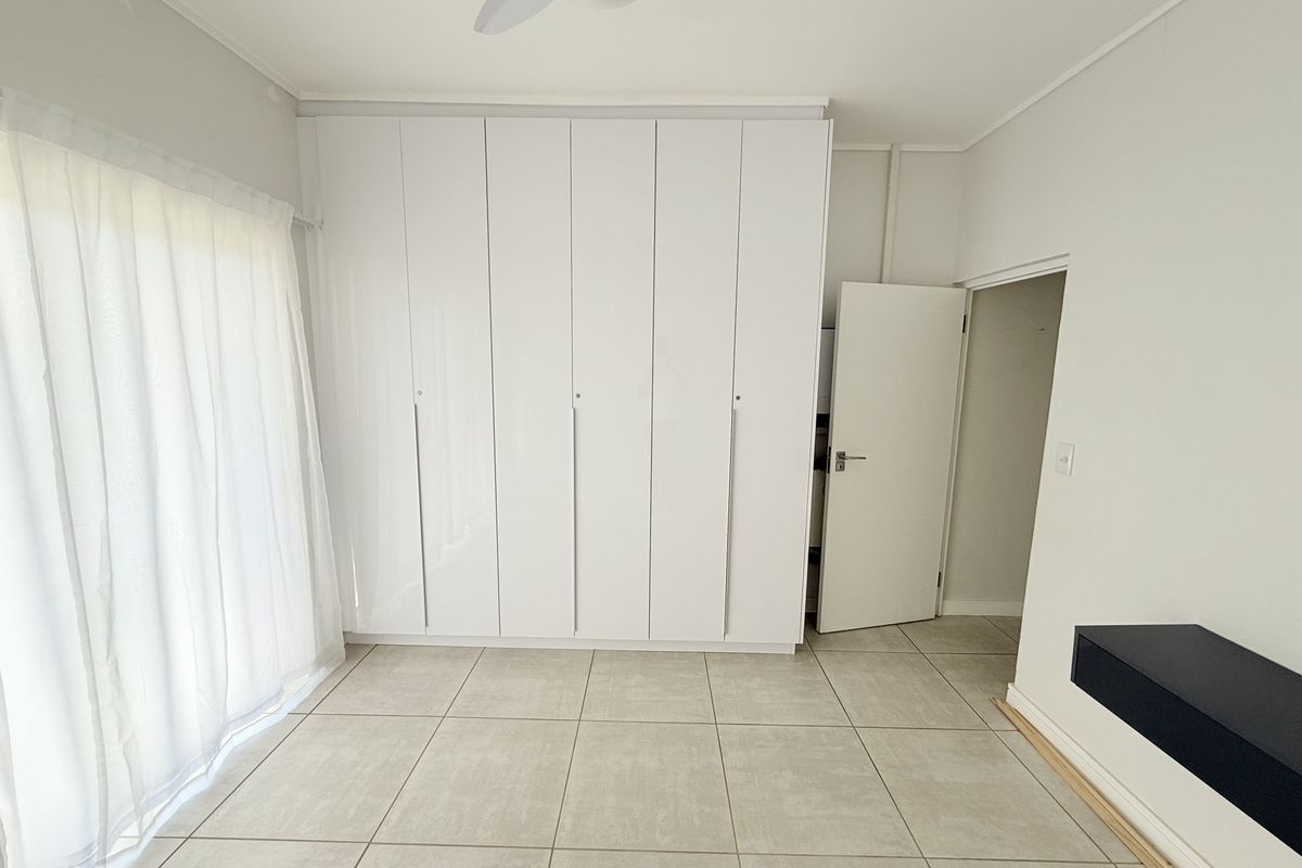 A bright, empty bedroom featuring neutral tiled flooring, extensive built-in white cupboards, a ceiling fan, and access to an outdoor area via a sliding door.