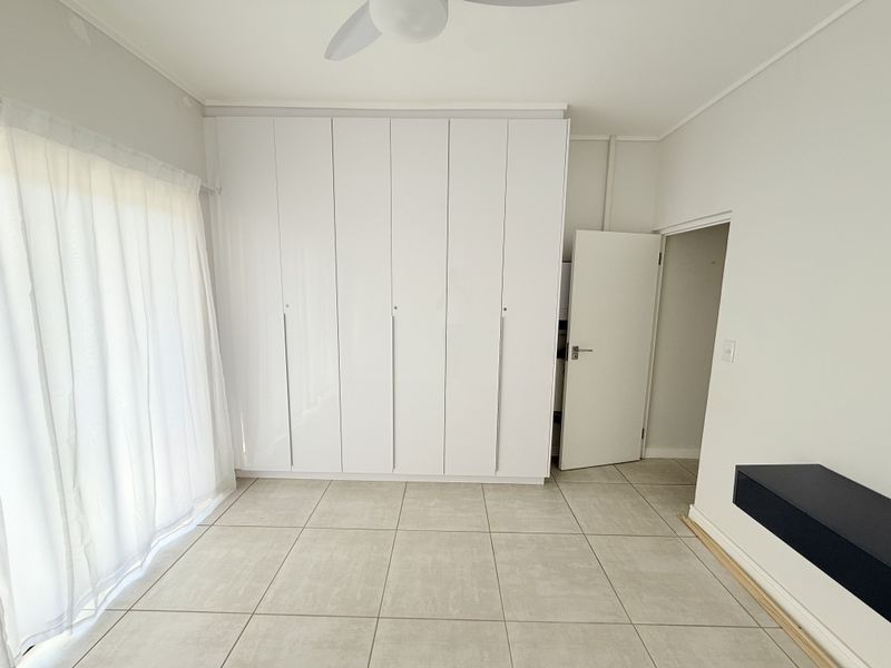 A bright, empty bedroom featuring neutral tiled flooring, extensive built-in white cupboards, a ceiling fan, and access to an outdoor area via a sliding door.