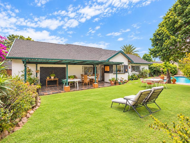 Rear view of the property featuring a covered patio, well-maintained lawn, and a swimming pool in a landscaped garden setting.