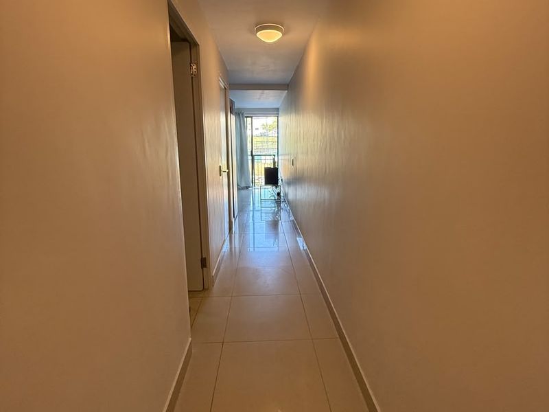 A clean, neutral-toned hallway featuring tiled flooring and a central ceiling light, leading towards an open living area with balcony access.
