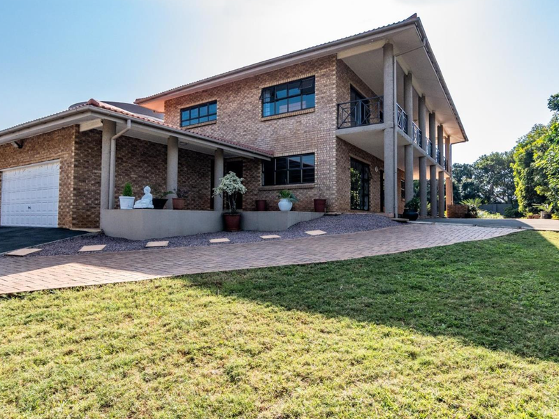 A well-maintained double-story brick house featuring a paved driveway, integrated garage, and a spacious covered balcony supported by concrete pillars.