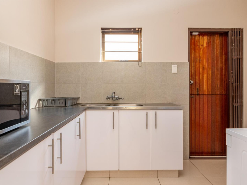 A clean and functional scullery area featuring white built-in cabinetry, a stainless steel sink, tiled splashbacks, and direct access to an exterior door.