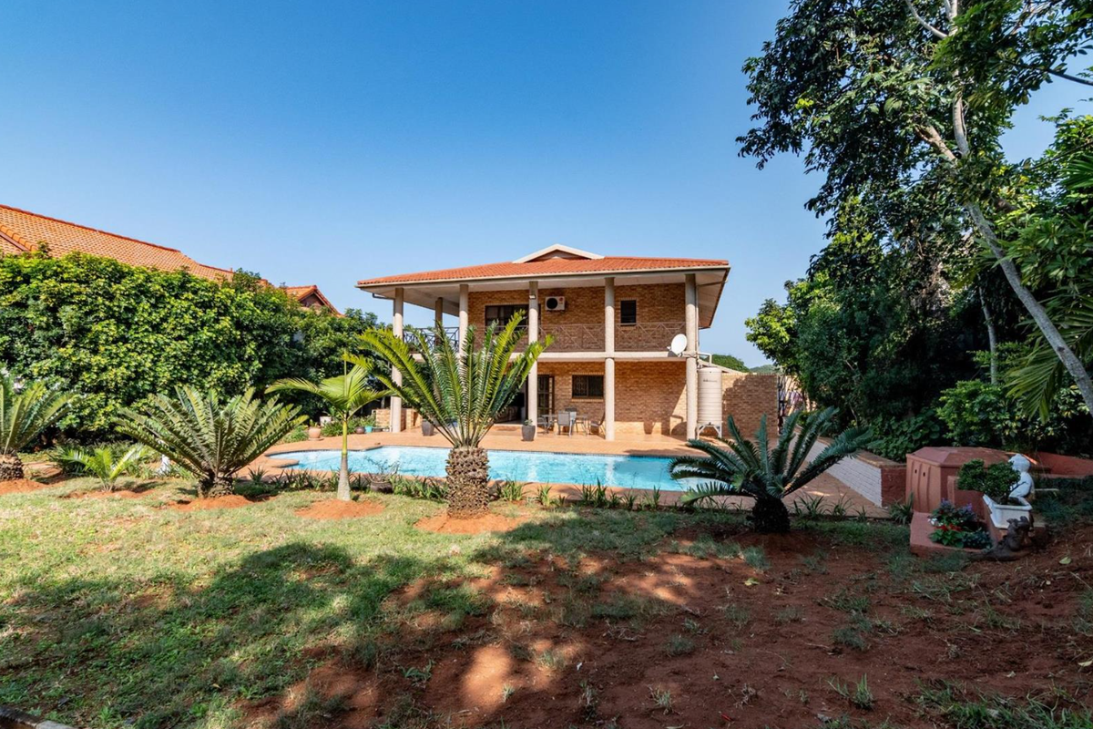 Rear view of a two-story brick house featuring a swimming pool, covered patio area, and established garden surroundings.