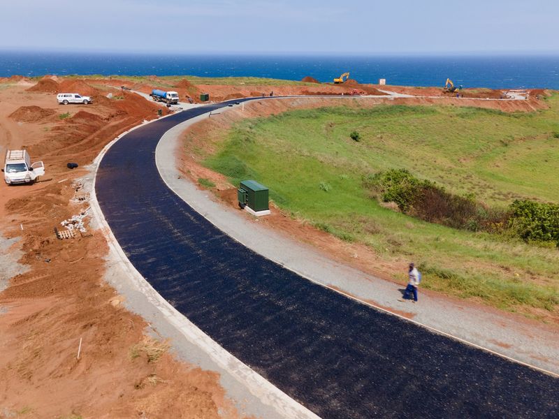 Aerial view of a newly paved asphalt road under construction within a residential development, featuring ocean views and surrounding cleared land.