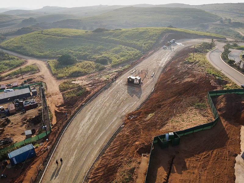Aerial view of a residential development site featuring an unpaved access road, security gatehouse, and perimeter fencing set within a hilly landscape.