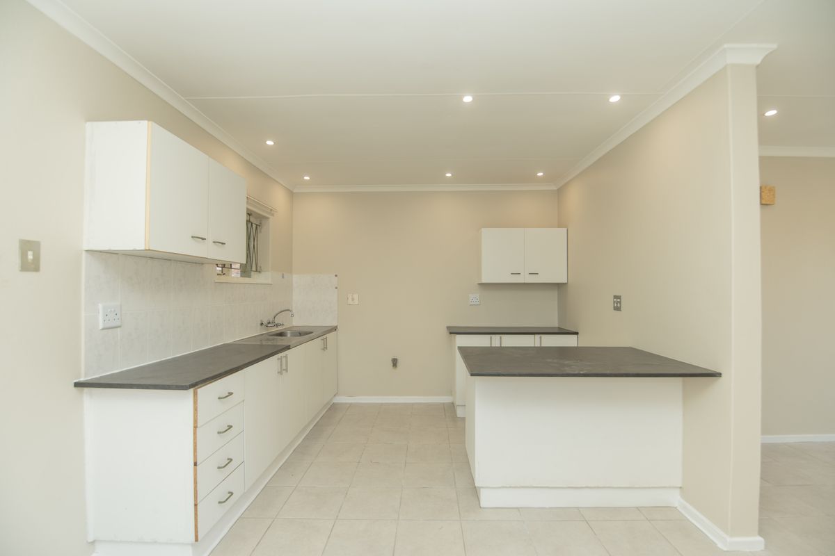 A functional, empty kitchen featuring white built-in cabinetry, dark laminate countertops, a stainless steel sink, and neutral tiled flooring with recessed lighting.