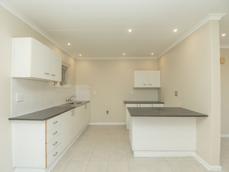 A functional, empty kitchen featuring white built-in cabinetry, dark laminate countertops, a stainless steel sink, and neutral tiled flooring with recessed lighting.