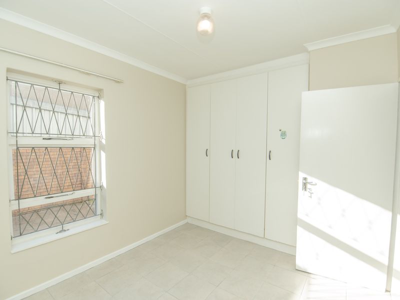 A bright, empty bedroom featuring neutral tiled flooring, built-in white cupboards, and a window fitted with security bars.