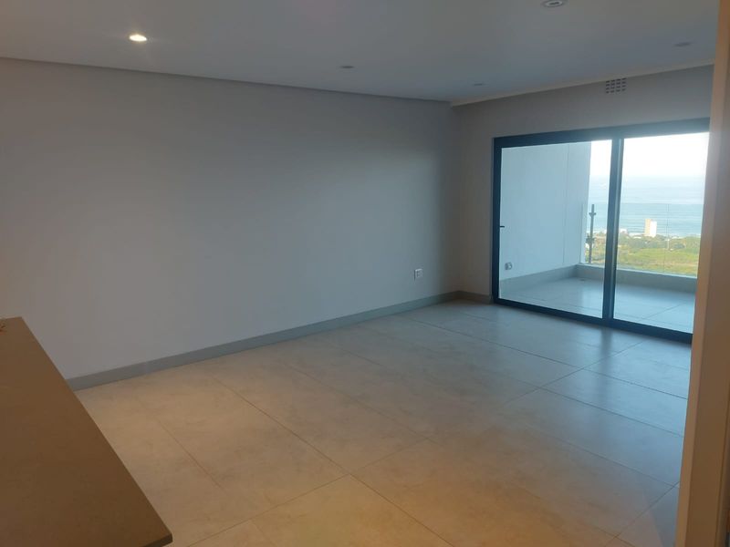 A bright, modern open-plan living area featuring large format floor tiles, recessed lighting, and sliding glass doors leading to a balcony with coastal views.