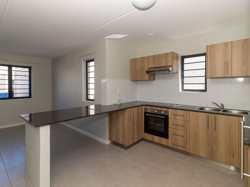 A clean, modern kitchen featuring wood-finish cabinetry, granite countertops, built-in oven and hob, and tiled flooring in an open-plan layout.
