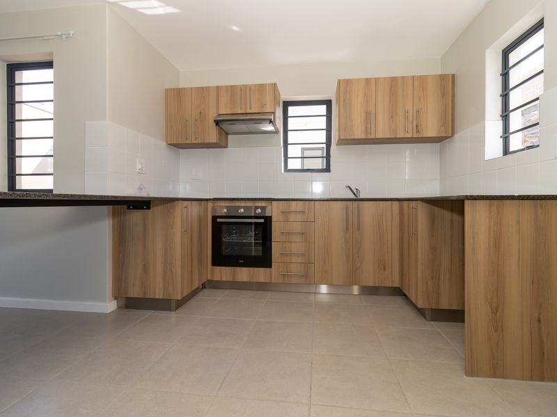 A clean, modern kitchen featuring wood-finish cabinetry, granite countertops, built-in oven, extractor fan, and tiled flooring with ample natural light.