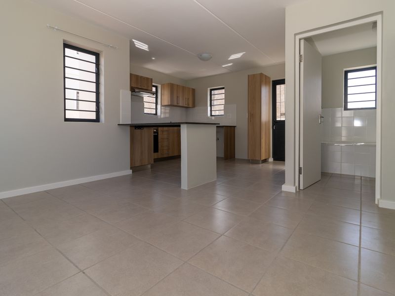 An open-plan kitchen and living area featuring neutral tiled flooring, modern wood-finish cabinetry, and a functional kitchen island, leading to an adjacent bathroom.