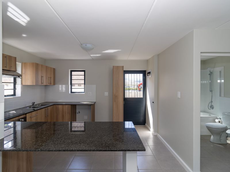 Modern open-plan kitchen featuring granite countertops, wood-finish cabinetry, and tiled flooring, with an adjacent bathroom and direct access to the exterior.