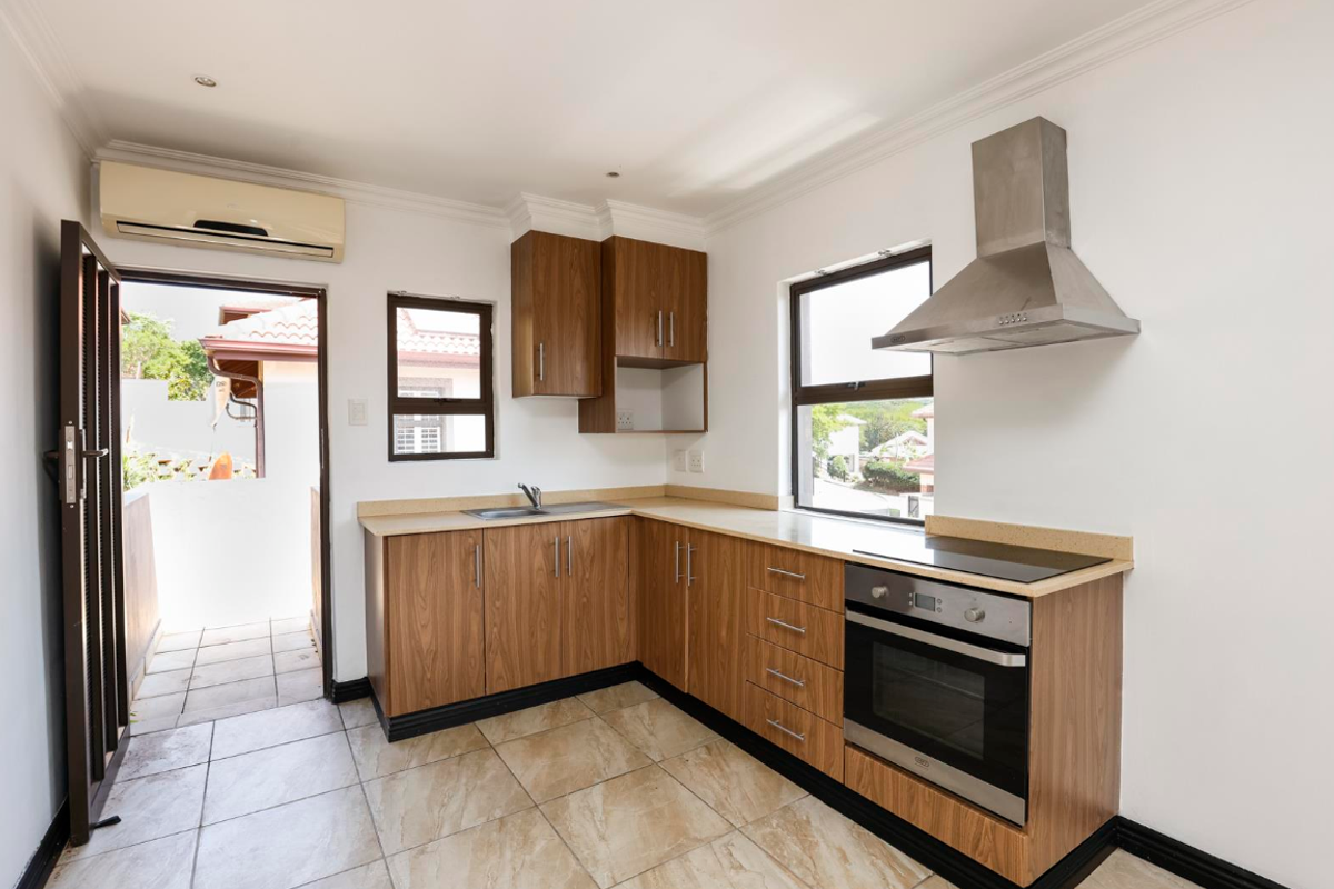 A clean, functional kitchen featuring wood-finish cabinetry, stone countertops, built-in oven, extractor fan, and direct access to an outdoor balcony area.
