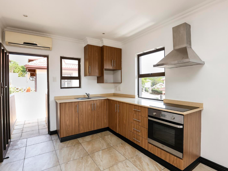 A clean, functional kitchen featuring wood-finish cabinetry, stone countertops, built-in oven, extractor fan, and direct access to an outdoor balcony area.
