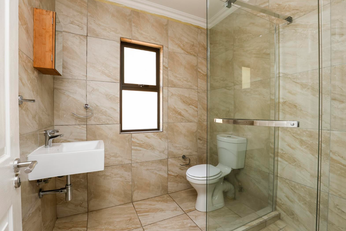 A modern bathroom featuring floor-to-ceiling beige marble-look tiles, a wall-mounted basin, a toilet, and a glass-enclosed shower.