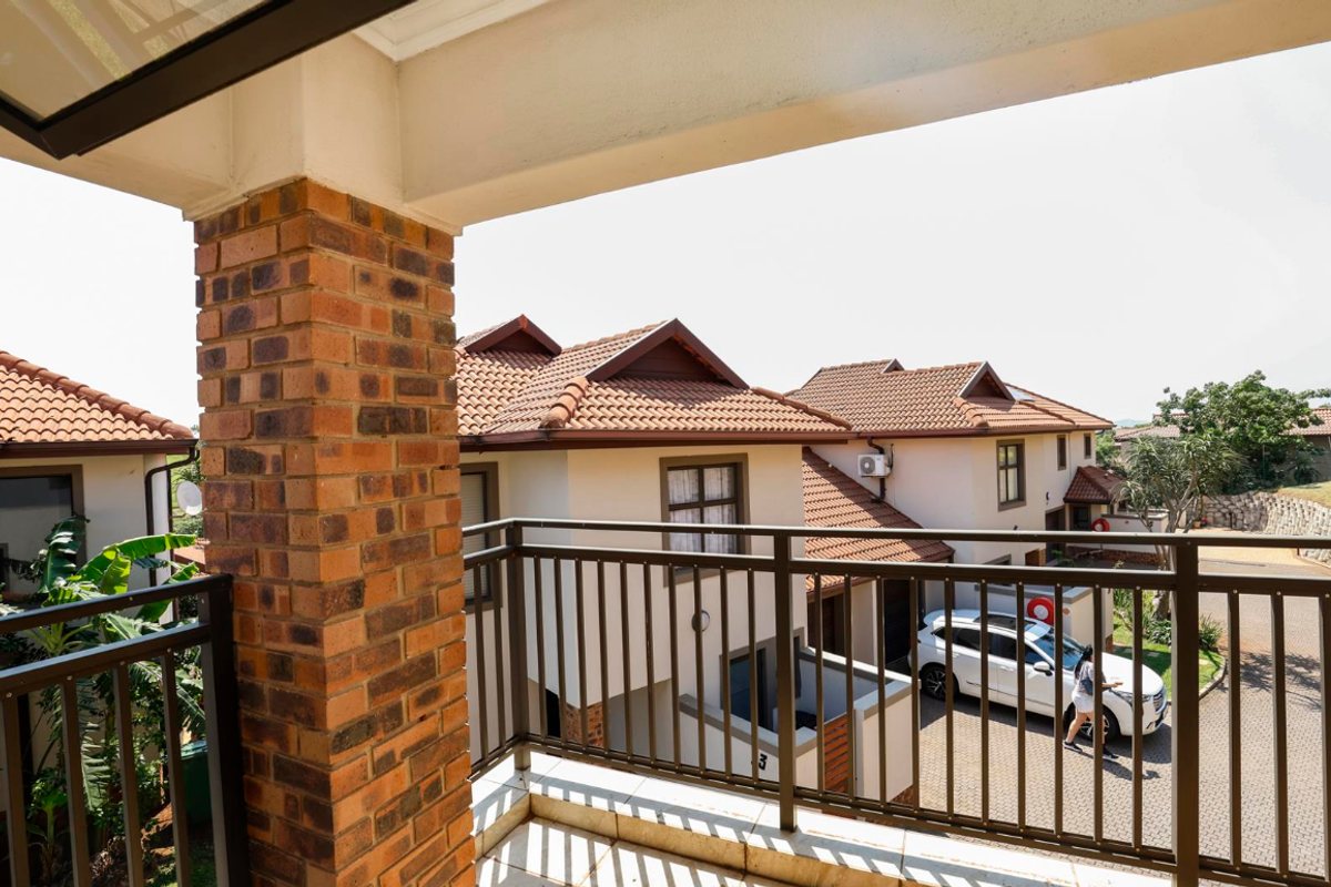 A covered balcony featuring brick pillar detailing, metal railings, and tiled flooring, overlooking the complex driveway and neighboring residential units.