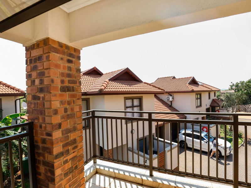A covered balcony featuring brick pillar detailing, metal railings, and tiled flooring, overlooking the complex driveway and neighboring residential units.