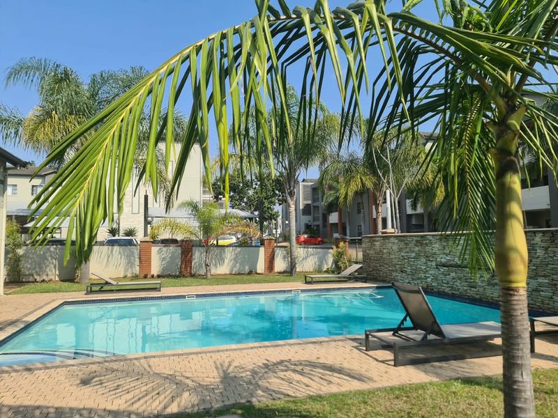 A well-maintained communal swimming pool area featuring stone cladding, paved surrounds, and established palm trees within a secure apartment complex.