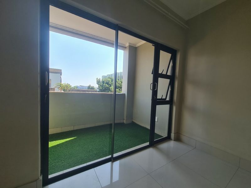 A bright, empty balcony featuring tiled flooring, artificial grass, and a large sliding glass door providing access from the interior.