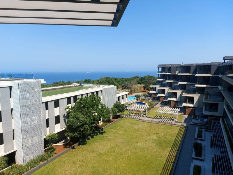 Elevated view overlooking the apartment complex grounds, featuring landscaped gardens, pergolas, a swimming pool, and a scenic ocean horizon.