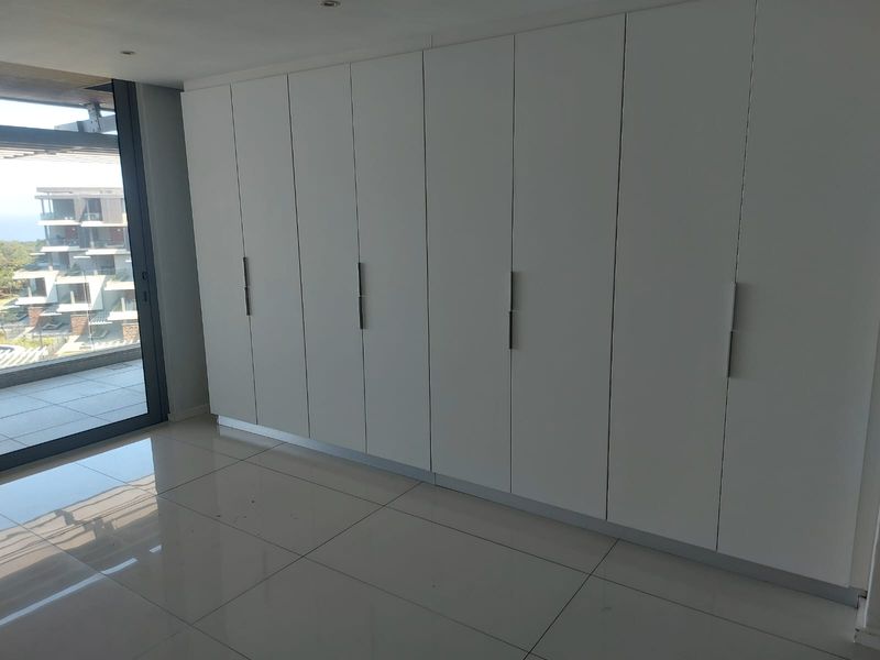 A bright bedroom featuring extensive built-in cupboards, sleek tiled flooring, and a large sliding glass door leading to an outdoor balcony area.