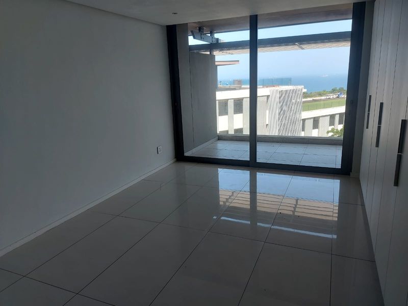 A bright, empty bedroom featuring modern tiled floors, built-in cupboards, and large sliding glass doors opening onto a balcony with ocean views.