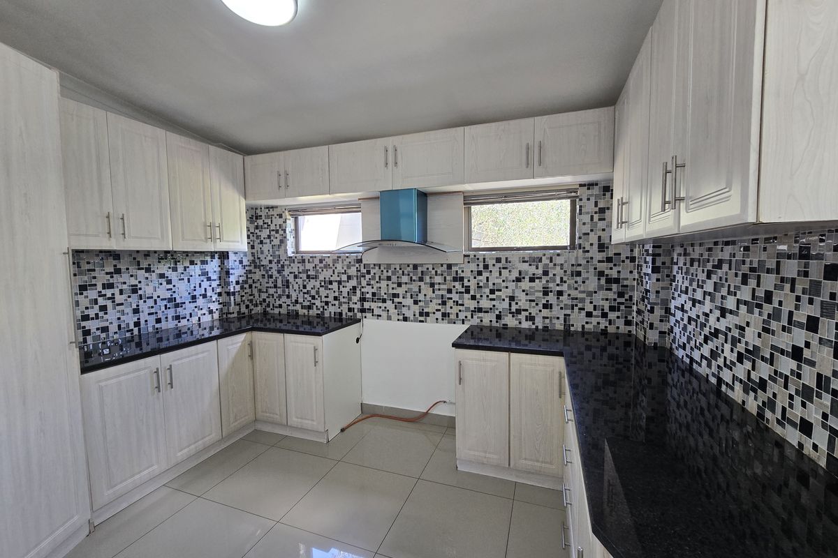 A clean, modern kitchen featuring light wood-finish cabinetry, contrasting black granite countertops, and a decorative mosaic tile backsplash with integrated extractor fan.