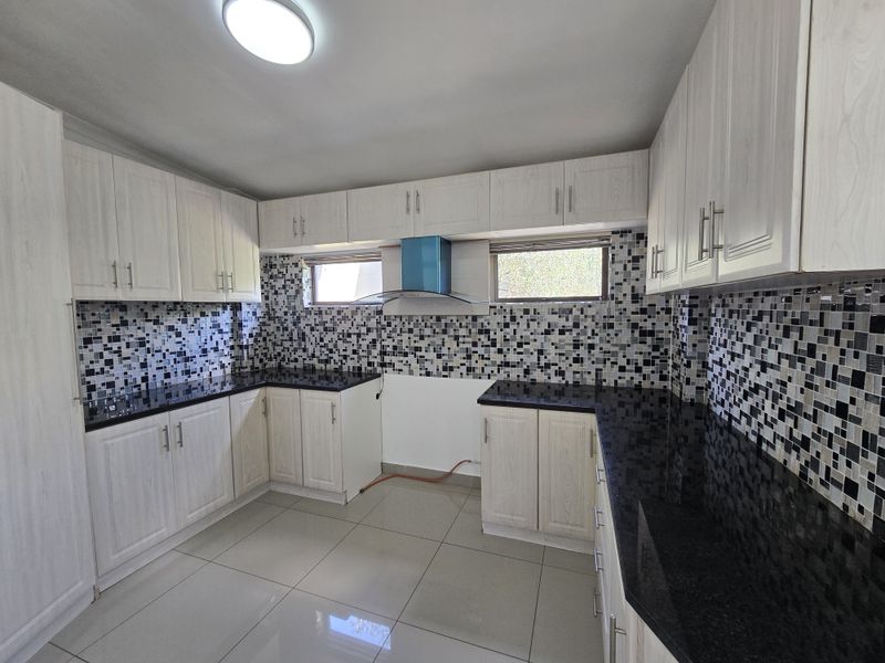 A clean, modern kitchen featuring light wood-finish cabinetry, contrasting black granite countertops, and a decorative mosaic tile backsplash with integrated extractor fan.