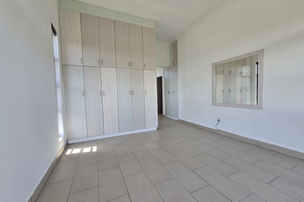 A bright, empty bedroom featuring neutral tiled flooring and extensive built-in cupboard storage with a matching wall-mounted mirror.