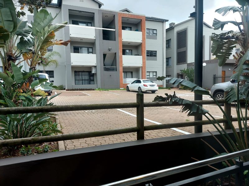 View of the apartment complex exterior featuring a paved parking area, multi-story building facade with balconies, and established landscaping.