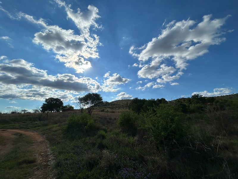 A scenic view of the expansive farm landscape featuring rolling hills, natural vegetation, and a dirt track under a bright, cloudy sky.
