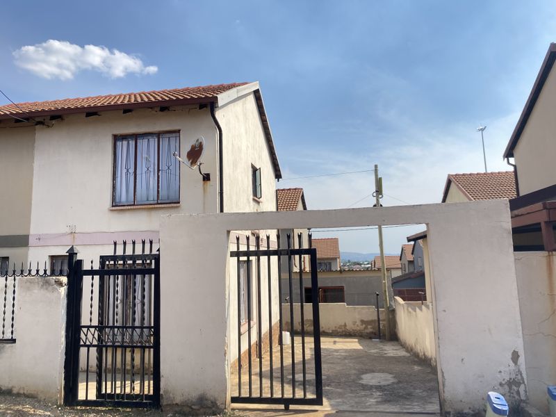 Front view of a two-story residential property featuring a concrete driveway, wrought iron security gate, and perimeter walling with visible exterior maintenance requirements.