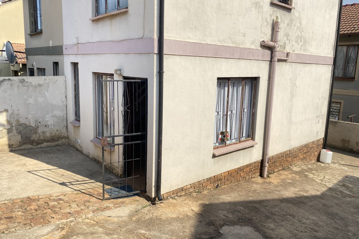 Rear exterior view of the property showing a paved courtyard, security gate, and weathered wall finishes requiring maintenance.