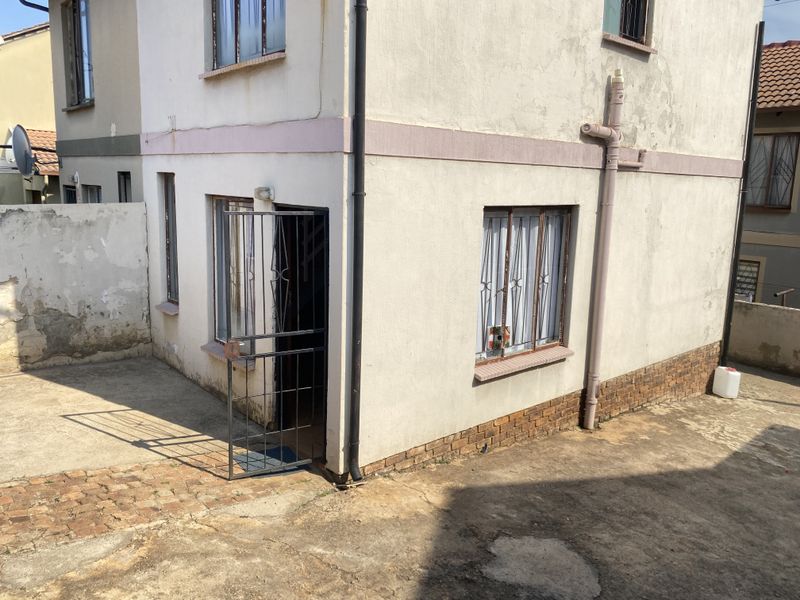 Rear exterior view of the property showing a paved courtyard, security gate, and weathered wall finishes requiring maintenance.