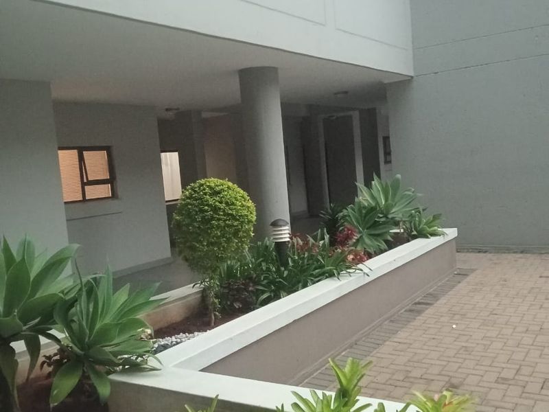 A well-maintained apartment complex courtyard featuring paved walkways, raised garden beds with established plants, and a covered structural walkway.